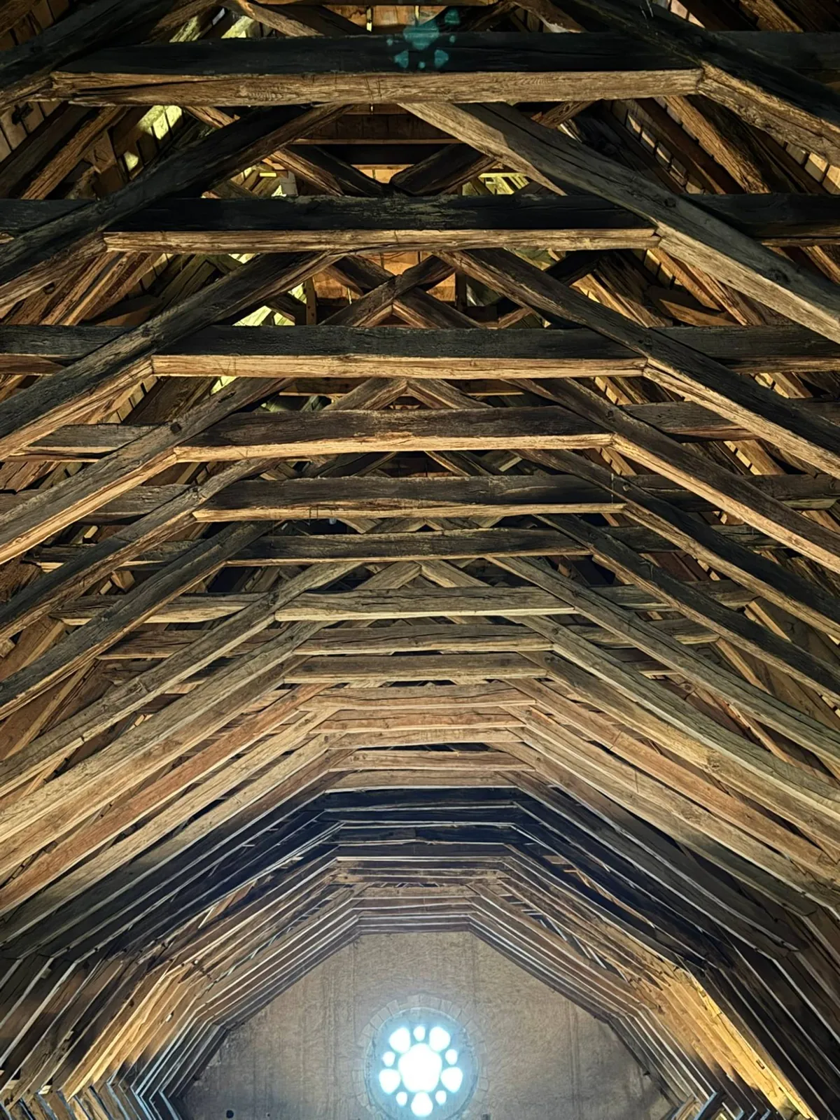 Charpente en bois des combles vue en enfilade, ses fermes à pans encadrant une petite fenêtre ronde au fond