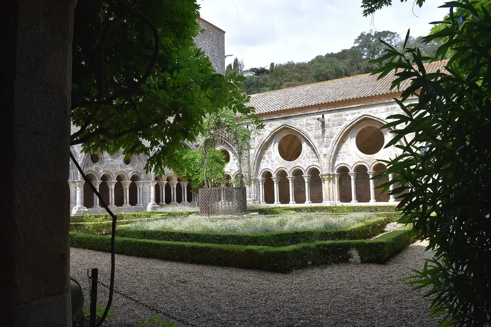 Cloître d'abbaye aux arcades de pierre, vu depuis une galerie ombragée, avec un jardin de buis au centre