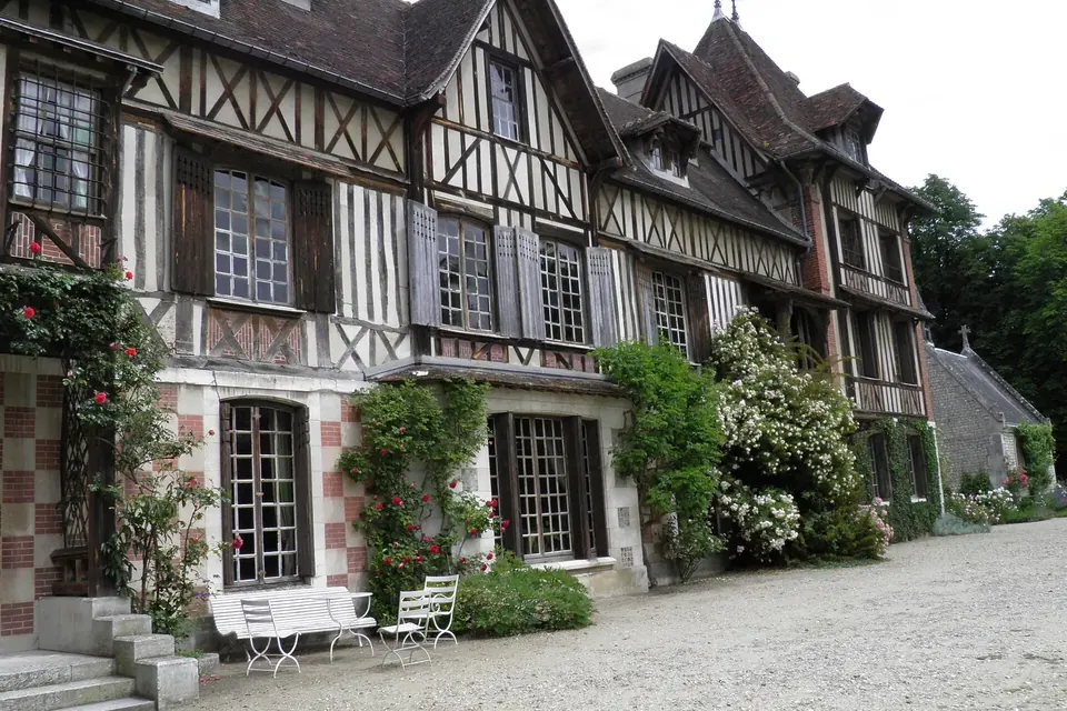 Façade de manoir normand à colombages et briques, fleurie de rosiers, avec bancs blancs devant l'entrée