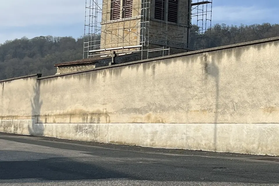 Long mur clair au bord d'une route, derrière lequel s'élève un clocher entouré d'échafaudages