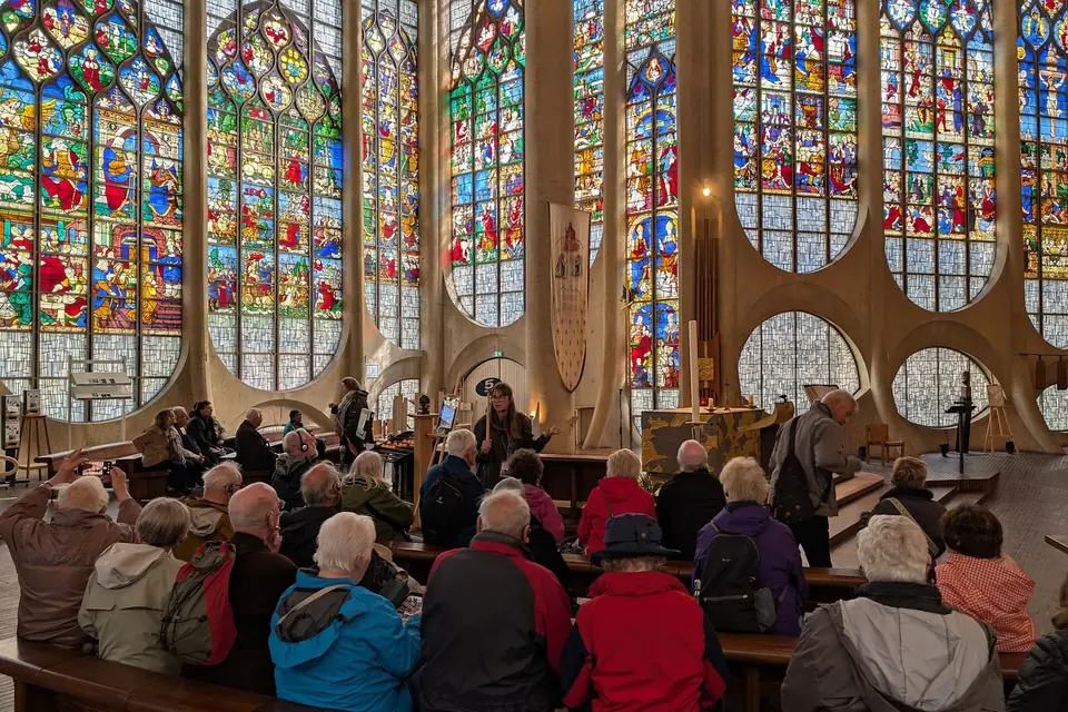 Groupe de visiteurs assis écoutant une guide devant de grands vitraux colorés